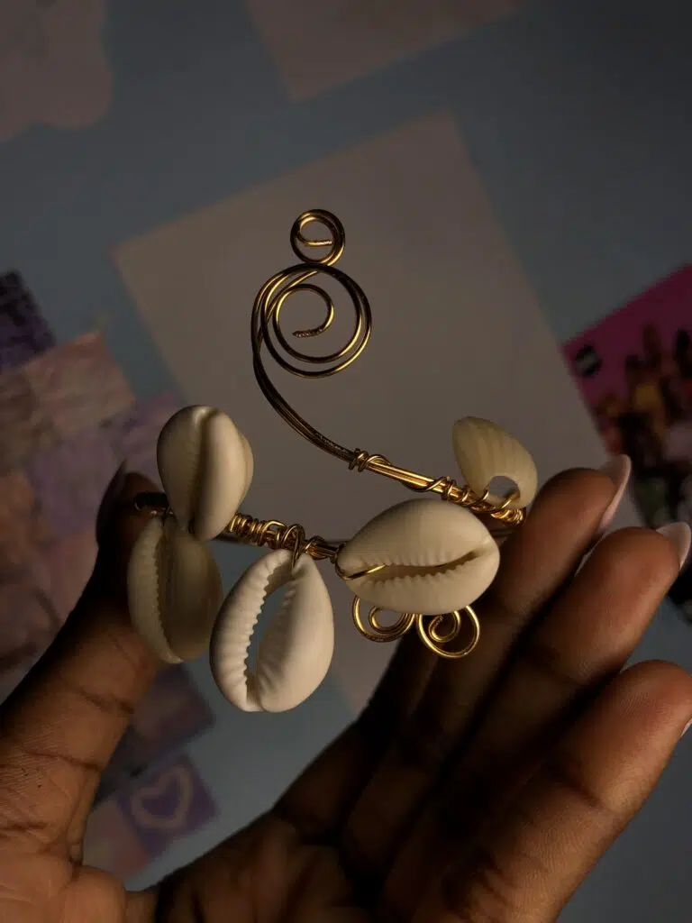 Gros plan sur un bijou de bras (brassard) artisanal sculptural tenu dans la main d'une femme à la peau foncée. Le bijou est formé d'un fil de métal doré épais, façonné à la main pour créer une structure rigide. De grands coquillages cauris blancs sont tissés et fixés au centre avec du fil de métal doré plus fin. Au sommet, le fil doré se déploie en une grande spirale élégante et un petit œil de métal. En bas, des volutes dorées encadrent les cauris. Le fond est sombre et légèrement flou.