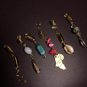 Gros plan sur sept bijoux de cheveux artisanaux, alignés en diagonale sur un fond noir mat. Chaque bijou commence par une bobine dorée torsadée destinée à se fixer sur une tresse. De cette bobine pendent des éléments uniques : Une perle tubulaire ocre et un serpentin doré. Une perle de quartz blanc lisse et un pendentif brut marron. Une perle tubulaire turquoise. Un chapelet de perles de corail rose et une plaque dorée en forme d'Afrique gravée avec « AFRICA IS GOOD ». Une perle en œil de tigre et une perle ovale blanche. Une perle cauri en argent et des perles de jade vert.