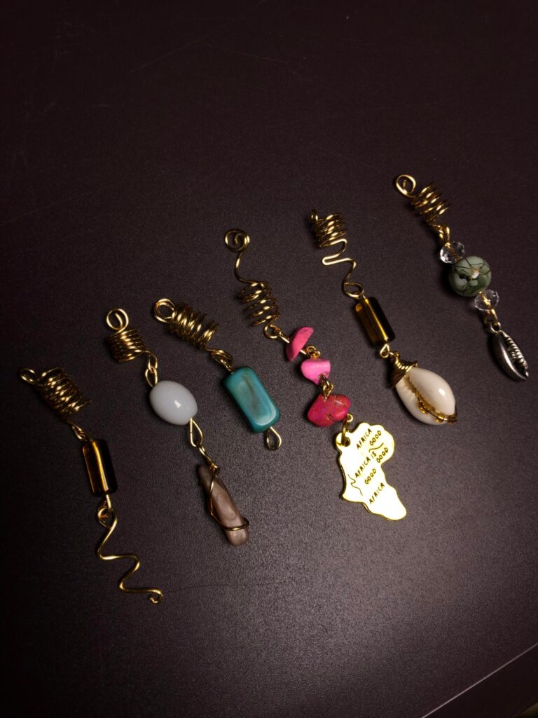 Gros plan sur sept bijoux de cheveux artisanaux, alignés en diagonale sur un fond noir mat. Chaque bijou commence par une bobine dorée torsadée destinée à se fixer sur une tresse. De cette bobine pendent des éléments uniques : Une perle tubulaire ocre et un serpentin doré. Une perle de quartz blanc lisse et un pendentif brut marron. Une perle tubulaire turquoise. Un chapelet de perles de corail rose et une plaque dorée en forme d'Afrique gravée avec « AFRICA IS GOOD ». Une perle en œil de tigre et une perle ovale blanche. Une perle cauri en argent et des perles de jade vert.
