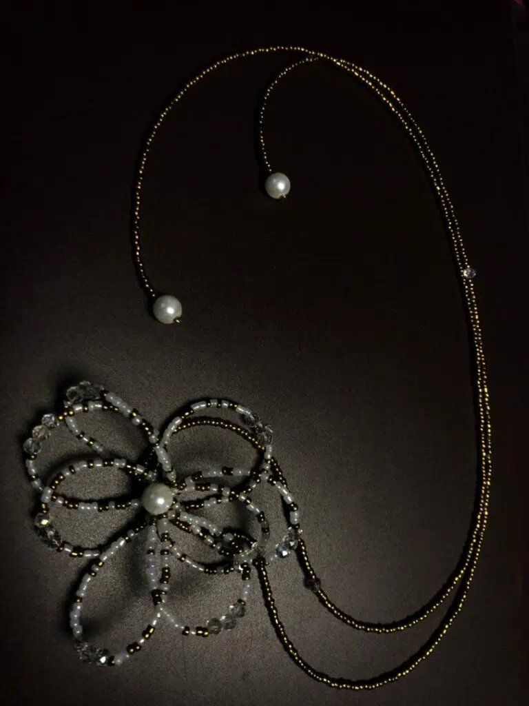 Gros plan sur un collier fantaisie en perles. Le bijou est formé d'une grande fleur stylisée en perles de rocaille blanches et transparentes, avec une perle nacrée au centre. De cette fleur part une fine chaîne de perles dorées qui se divise en deux ornant le cou et plongeant en cravate avec une perle blanche à aux extrémités.