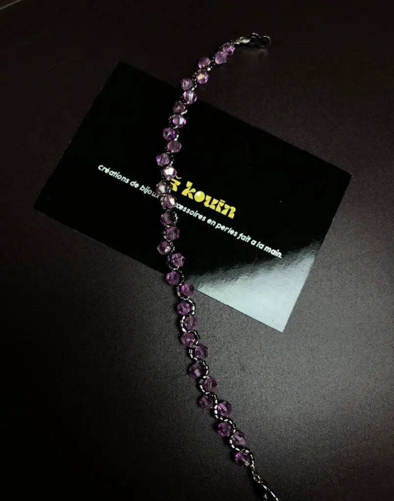Un bracelet artisanal fin posé en diagonale sur un fond sombre. Il est composé de perles de cristal facettées de couleur lilas/mauve, tissées en zigzag avec de minuscules perles de rocaille argentées. Le bracelet traverse une carte de visite noire où l'on peut lire en lettres dorées : « Jekouin - Créations de bijoux et accessoires en perles fait à la main ». Un fermoir argenté est visible à l'extrémité.