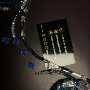 Un bijou de taille (baya) artisanal disposé en boucles sur un fond sombre. Il est composé de perles de rocaille bleu roi mates et de perles de cristal facettées transparentes, alternant avec des perles en tube allongées et transparentes. Une fine chaîne dorée suspendue comporte quatre motifs de trèfles à quatre feuilles émaillés : trois en bleu vif et un en blanc perlé, tous encadrés d'un bord perlé doré.