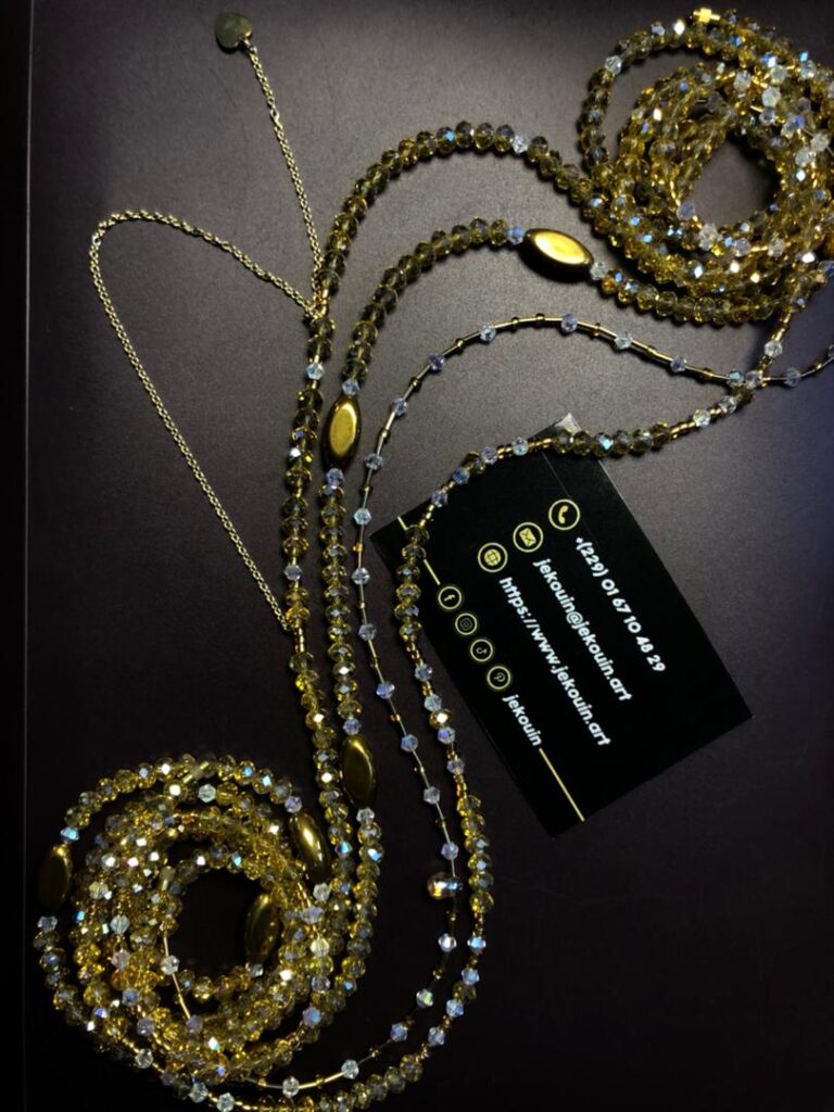Un bijou de taille (baya) artisanal de couleur dorée et ambrée, disposé en boucles fluides sur un fond noir. Il est composé de perles de cristal facettées qui scintillent, entrecoupées de perles ovales lisses de couleur bronze doré. Une fine chaîne dorée avec un petit pendentif en forme de cœur termine l'ensemble. La carte de visite noire de Jekouin.art est placée à côté du bijou.