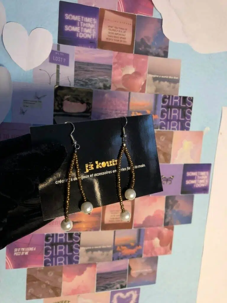 Une main gantée de noir présente une carte de visite noire supportant une paire de boucles d'oreilles pendantes artisanales de la marque Jekouin.art. Chaque boucle est composée d'un mince fil de perles de rocaille dorées qui serpente légèrement, orné de deux perles nacrées blanches : une au centre et une à l'extrémité. Le fond est un mur décoré d'un collage de photos esthétiques aux tons violet, rose et bleu, avec des inscriptions comme « GIRLS GIRLS GIRLS » et des motifs de cœurs et papillons.