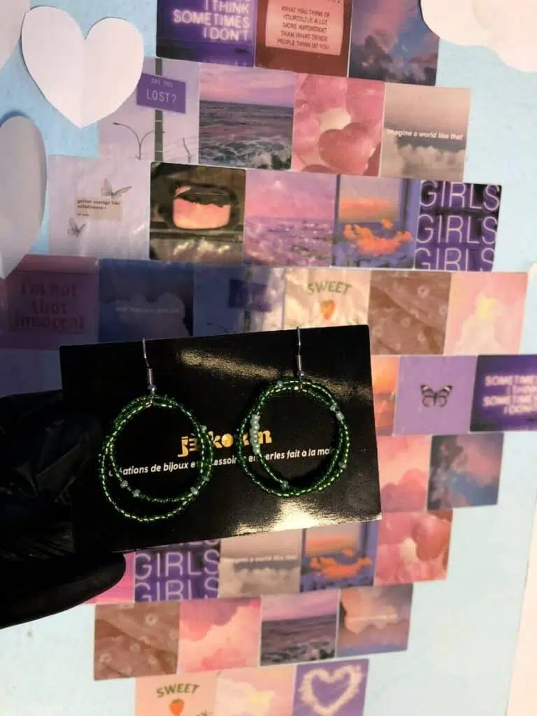 Une paire de boucles d'oreilles créoles artisanales présentées sur une carte de visite noire de la marque Jekouin.art. Chaque boucle est formée de deux cercles entrelacés : l'un plus grand en perles de rocaille vert émeraude scintillantes, et l'un plus petit avec des touches de perles vert menthe pastel. Le bijou est tenu devant un mur décoré d'un collage de photos aux tons violets et rosés. Les crochets sont de couleur argentée.