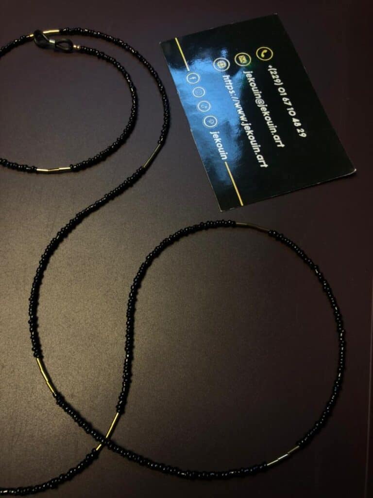 Gros plan d'une chaîne de lunettes artisanale délicate. La chaîne est composée de minuscules perles de rocaille noires brillantes, ponctuées de segments de perles intercalaires dorées plus longues. Elle est munie d'attaches élastiques noires avec des anneaux dorés à chaque extrémité. La chaîne est disposée sur un fond sombre et uni, avec une carte de visite noire de la marque Jekouin.art visible en arrière-plan.