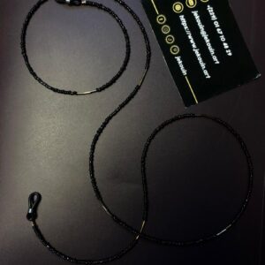 Gros plan d'une chaîne de lunettes artisanale délicate. La chaîne est composée de minuscules perles de rocaille noires brillantes, ponctuées de segments de perles intercalaires dorées plus longues. Elle est munie d'attaches élastiques noires avec des anneaux dorés à chaque extrémité. La chaîne est disposée sur un fond sombre et uni, avec une carte de visite noire de la marque Jekouin.art visible en arrière-plan.