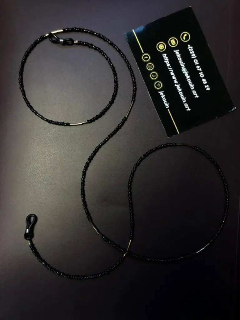 Gros plan d'une chaîne de lunettes artisanale délicate. La chaîne est composée de minuscules perles de rocaille noires brillantes, ponctuées de segments de perles intercalaires dorées plus longues. Elle est munie d'attaches élastiques noires avec des anneaux dorés à chaque extrémité. La chaîne est disposée sur un fond sombre et uni, avec une carte de visite noire de la marque Jekouin.art visible en arrière-plan.