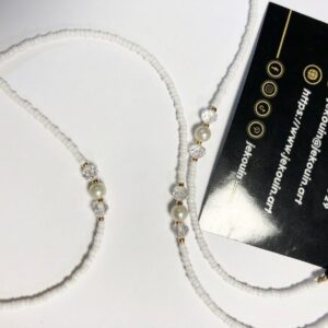 Gros plan sur une chaîne de lunettes artisanale posée en boucle sur un fond blanc neutre. La chaîne est principalement composée de minuscules perles de rocaille blanches mates. À intervalles réguliers, elle est ornée de perles plus grandes : une perle nacrée blanche ronde encadrée par deux cristaux facettés transparents, le tout sécurisé par de petites perles intercalaires dorées. À une extrémité, on voit des attaches élastiques noires avec des anneaux dorés. Une carte de visite noire avec des logos de contact en or et les informations « Jekouin » est visible dans le coin supérieur droit.