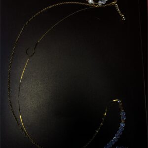 Bijou de taille (baya) composé d'une fine chaîne métallique centrale entourée de perles de verre transparentes captant la lumière, avec des reflets rouges, jaunes et bleus selon l'inclinaison, de perles orangées et de perles blanches.