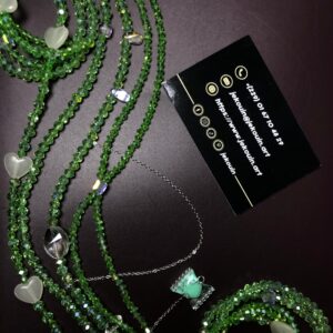 Baya (bijou de taille) composé de plusieurs rangs de perles dans un dégradé de verts : vert forêt, vert menthe et touches de blanc crème, assemblé sur un fil résistant. Une carte de visite noire de « Jekouin.art » avec logos dorés est placée à coté.