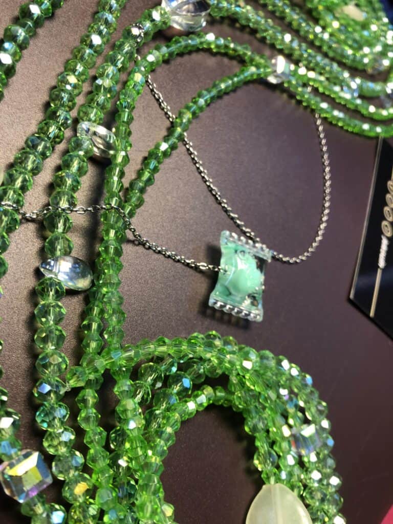 Baya (bijou de taille) composé de plusieurs rangs de perles dans un dégradé de verts : vert forêt, vert menthe et touches de blanc crème, assemblé sur un fil résistant. Focus sur un pendentif en forme de bonbon.