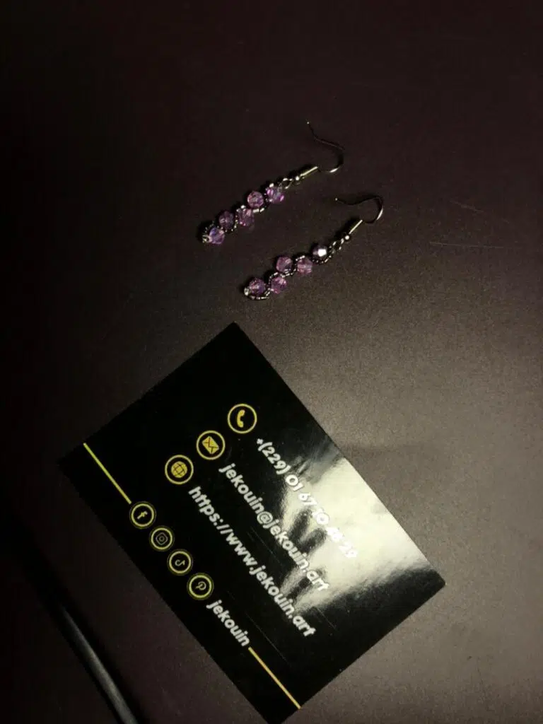 Paire de boucles d'oreilles pendantes composées de perles de cristal facettées de couleur mauve/lilas, articulées en zigzag sur une fine structure argentée. Elles sont présentées à côté d'une carte de visite noire Jekouin.art sur un fond sombre.