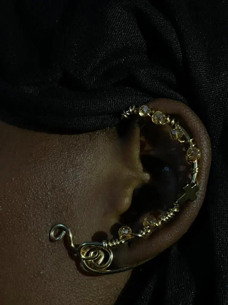 Gros plan sur une oreille humaine à la peau foncée portant un earcuff artisanal en fil de métal doré. Le bijou épouse la forme de l'hélix et du lobe. Il présente une rangée de perles facettées ambrées, une petite croix dorée au centre et se termine en bas par une spirale de métal plus épaisse reposant sur le lobe. Le fond est un drapé de tissu noir sombre.