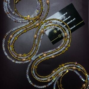 Deux longs bijoux de taille (bayas) entrelacés sur un fond noir. Le premier est composé de perles dorées scintillantes rythmées par des perles nacrées ovales et de petites croix dorées. Le second est constitué de perles de cristal facettées aux reflets irisés (AB) parsemées de touches jaunes. La carte de visite de Jekouin.art est placée au centre de la composition.