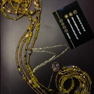 Un bijou de taille (baya) artisanal de couleur dorée, disposé de manière fluide sur un fond sombre. Il est composé de multiples rangs de perles de rocaille dorées, de perles facettées jaunes transparentes, de perles nacrées ovales et de perles tubes en verre. Une fine chaîne en argent de style maille Figaro s'entrelace avec les perles. Un pendentif transparent en forme de coquillage est suspendu au centre. La carte de visite Jekouin.art accompagne l'ensemble.