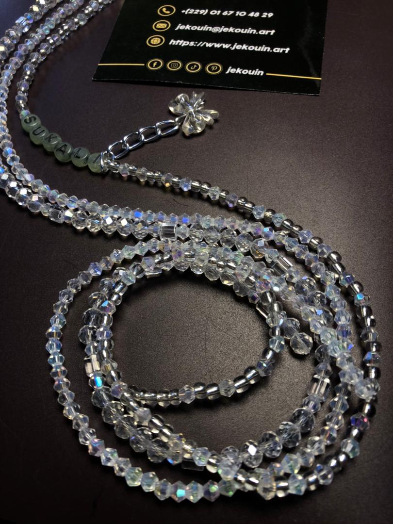 Baya (chaîne de taille) en perles de cristal facettées transparentes aux reflets irisés, disposé sur un fond sombre. Une section comporte des perles plates grises formant le nom « SUKALI ». Une petite chaîne argentée avec une breloque en forme de fleur est visible en haut, à côté d'une carte de visite noire de Jekouin.art.