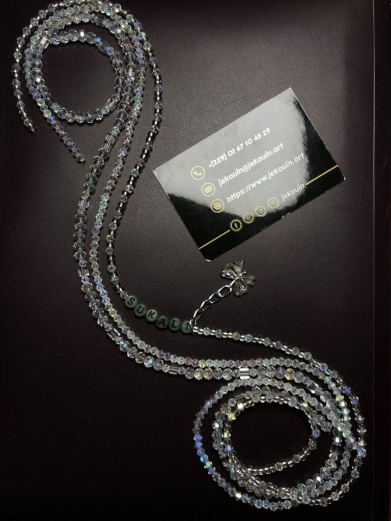 Baya (chaîne de taille) en perles de cristal facettées transparentes aux reflets irisés, disposé sur un fond sombre. Une section comporte des perles plates grises formant le nom « SUKALI ». Une petite chaîne argentée avec une breloque en forme de fleur est visible en haut, à côté d'une carte de visite noire de Jekouin.art.