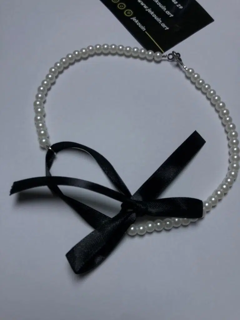 Collier de perles blanches rondes et uniformes, disposé en demi-cercle sur un fond sombre. Un ruban noir satiné est noué à l'avant du collier. Une carte de visite noire de « Jekouin.art » avec logos dorés est placée en dessous.