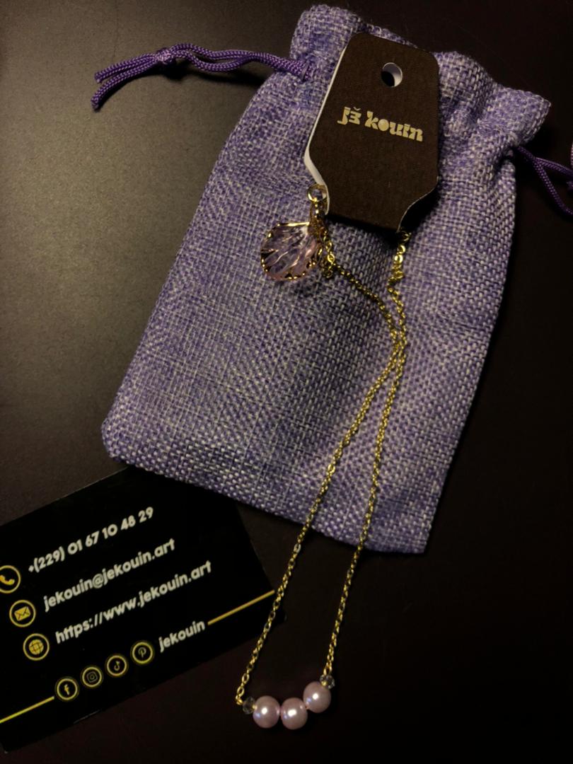 Collier fin avec une chaîne dorée présentant deux pendentifs distincts de couleur violette, conçus pour être interchangeables. L'un est un pendentif violet en forme de coquillage, l'autre est un pendentif violet constitué de trois perles nacrées assorties. Une carte de visite noire de « Jekouin.art » avec logos dorés est placée en dessous.