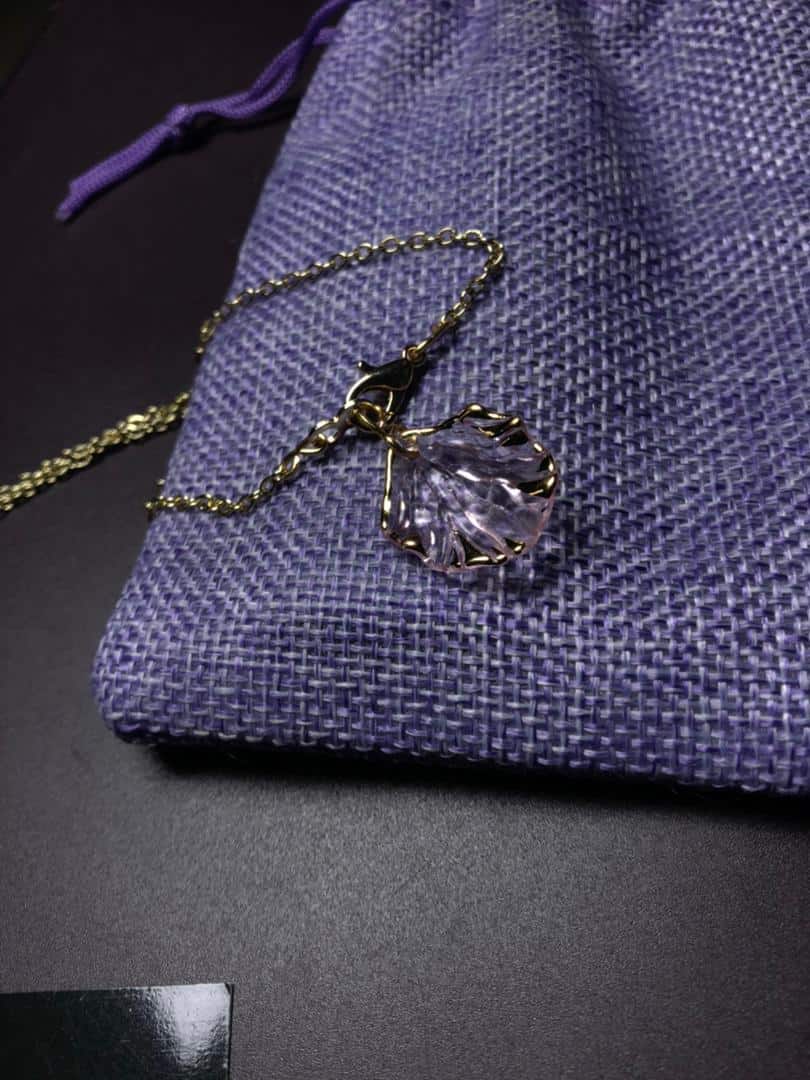 Collier fin avec une chaîne dorée présentant deux pendentifs distincts de couleur violette, conçus pour être interchangeables. L'un est un pendentif violet en forme de coquillage, l'autre est un pendentif violet constitué de trois perles nacrées assorties. Focus sur le pendentif coquillage.
