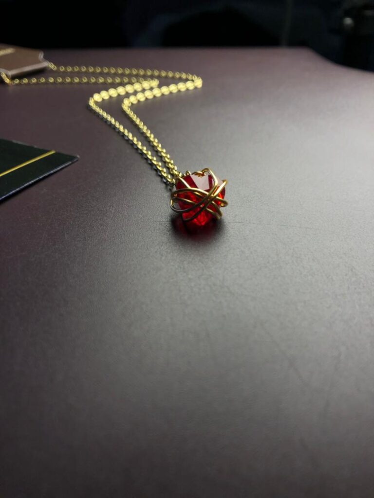 Collier avec un pendentif composé d'une pierre rouge sculptée, qui est fermement maintenue par un enroulement complexe et décoratif de fil doré formant une monture artisanale unique.