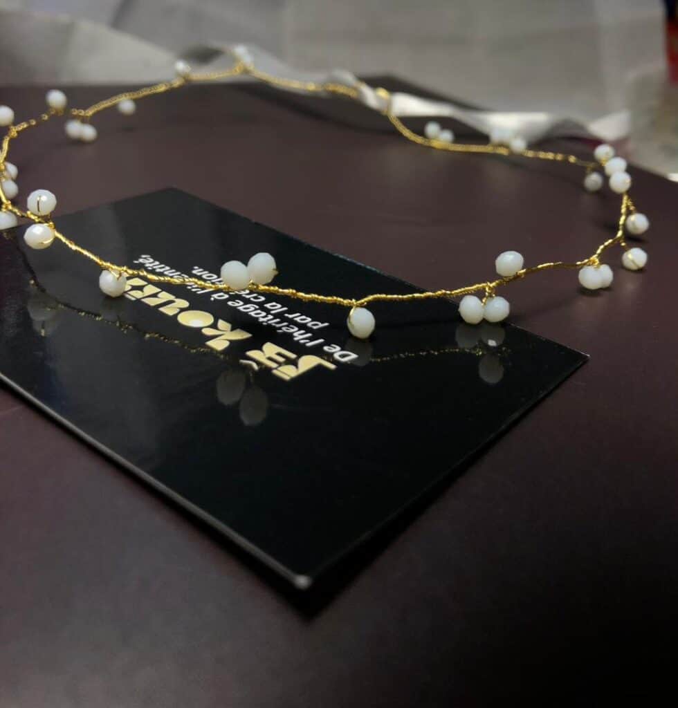 Vue rapprochée d'un diadème délicat en fil d'or torsadé, orné de petites perles blanches, reposant sur une surface sombre. Une carte de visite noire de la marque "jǯ kouin" est visible au premier plan, avec un ruban de satin blanc en arrière-plan.