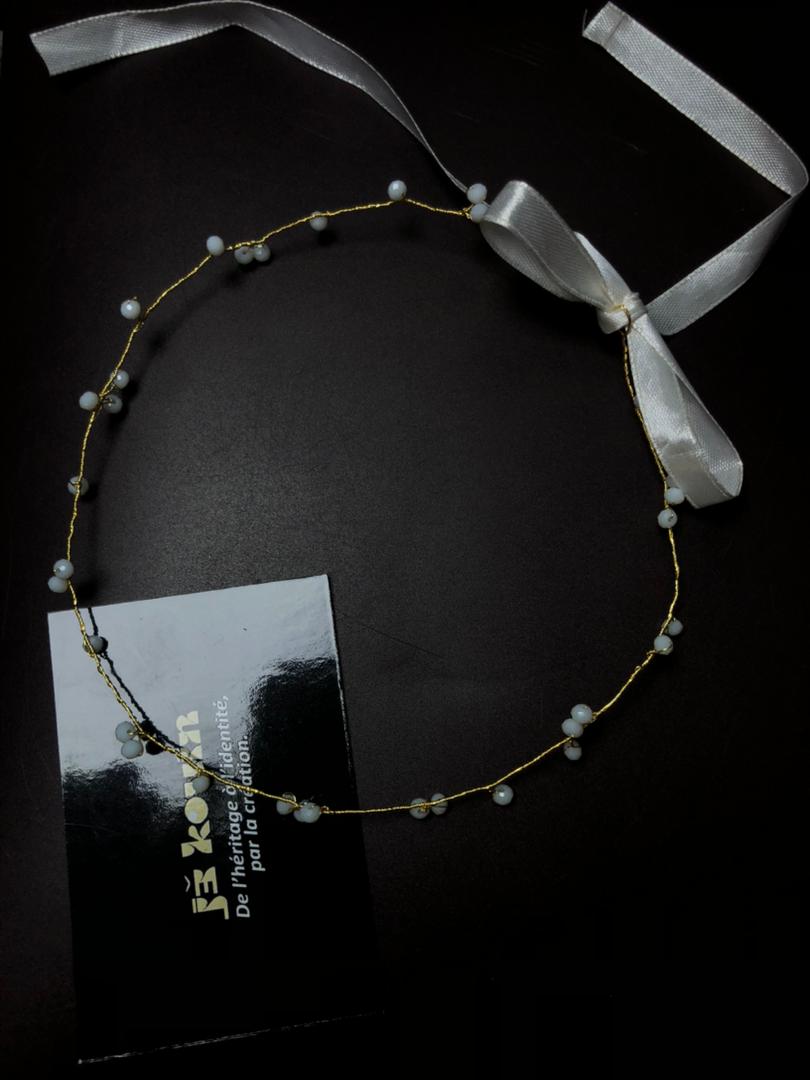 Photographie prise d'en haut d'un diadème délicat en fil d'or et perles de lait, formant un cercle imparfait sur une surface noire mate. Une carte de visite jǯ kouin est placée en bas à gauche et un ruban de satin blanc avec un nœud est en haut à droite.
