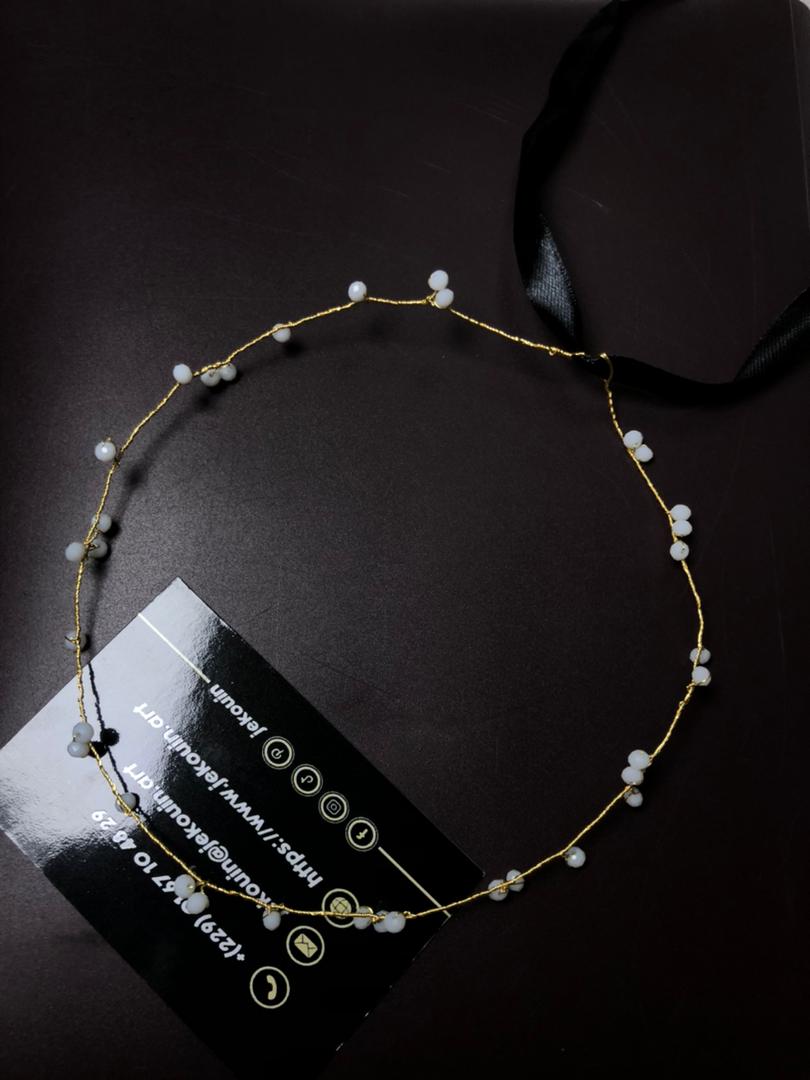 Vue d'ensemble d'un diadème délicat en fil d'or et perles de lait, disposé en cercle imparfait sur une surface noire mate. Une carte de visite jǯ kouin avec les coordonnées est en bas à gauche, et un ruban de satin noir s'étire vers le haut à droite.