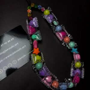 Un accessoire de téléphone coloré composé de perles en forme de bonbons enveloppés, d'étoile et de formes pastel, formant une boucle sur fond noir à côté d'une carte jǯ kouin.