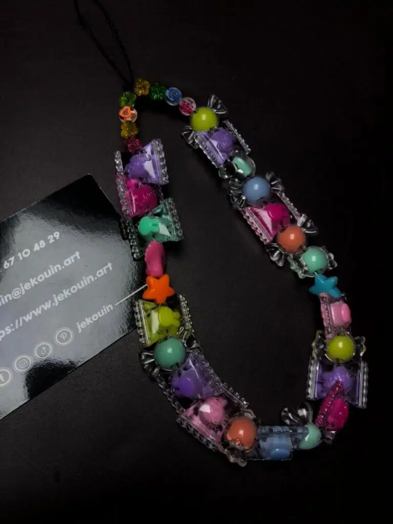 Un accessoire de téléphone coloré composé de perles en forme de bonbons enveloppés, d'étoile et de formes pastel, formant une boucle sur fond noir à côté d'une carte jǯ kouin.