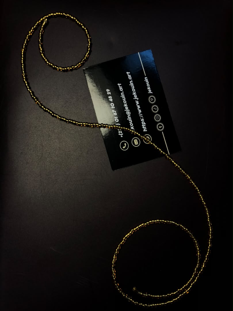 Un unique fil de taille en perles fines de rocaille dorées, disposé en arabesque autour d'une carte de visite jǯ kouin sur un fond noir texturé.
