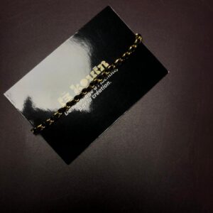 Un bracelet fin composé d'une chaîne dorée entrelacée de fil noir, disposé en diagonale sur la carte de visite noire jǯ kouin.