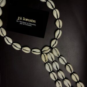 Collier en coquillages cauris blancs montés sur fil noir avec une section principale circulaire et trois longs rangs pendants, posé avec la carte jǯ kouin.