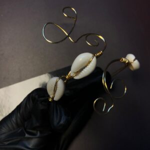 Création artisanale en fil métallique doré façonnée en volutes et volubiles ornée de coquillages cauris blancs, présentée par une main gantée de noir.