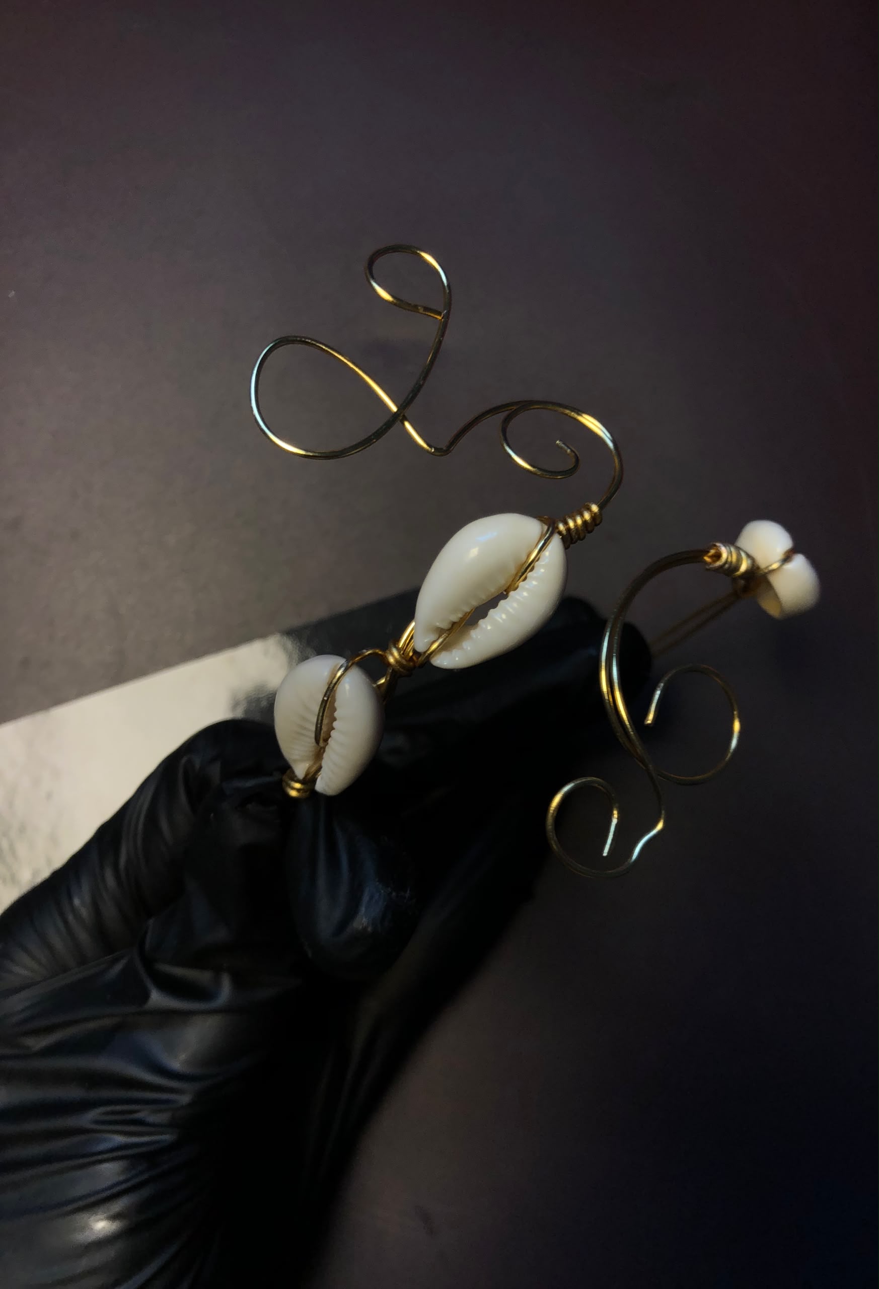 Création artisanale en fil métallique doré façonnée en volutes et volubiles ornée de coquillages cauris blancs, présentée par une main gantée de noir.