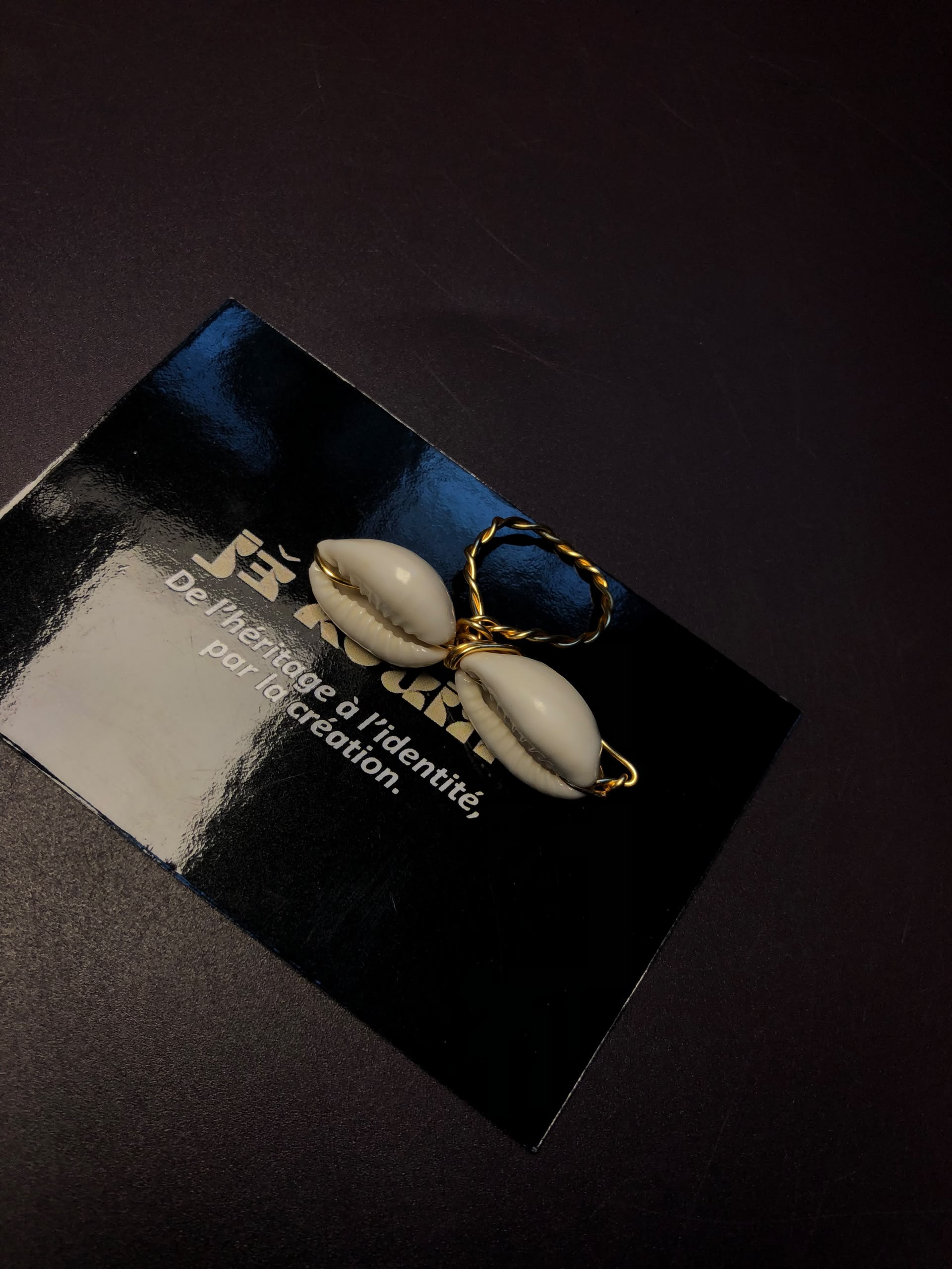 Gros plan sur une bague composée de deux coquillages cauris blancs polis, montés sur un anneau en fil d'or torsadé, posée sur une carte jekouin noire.