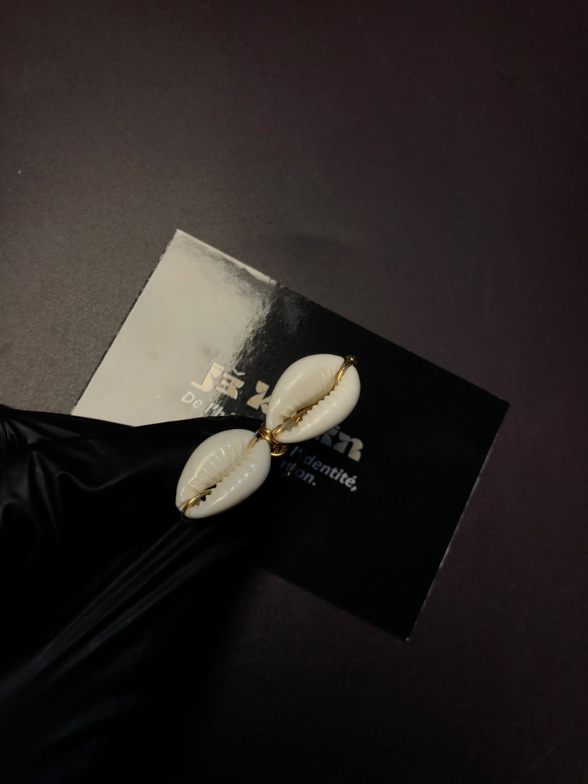 Vue d'ensemble montrant une bague double cauris avec un anneau en fil d'or torsadé, portée au doigt d'une main gantée de noir, à côté de la carte jekouin.