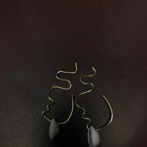 Paire de boucles d'oreilles originales en fil d'or façonnées à la main dessinant le profil abstrait d'un visage avec un cauris naturel suspendu à la base, posées sur un fond sombre.