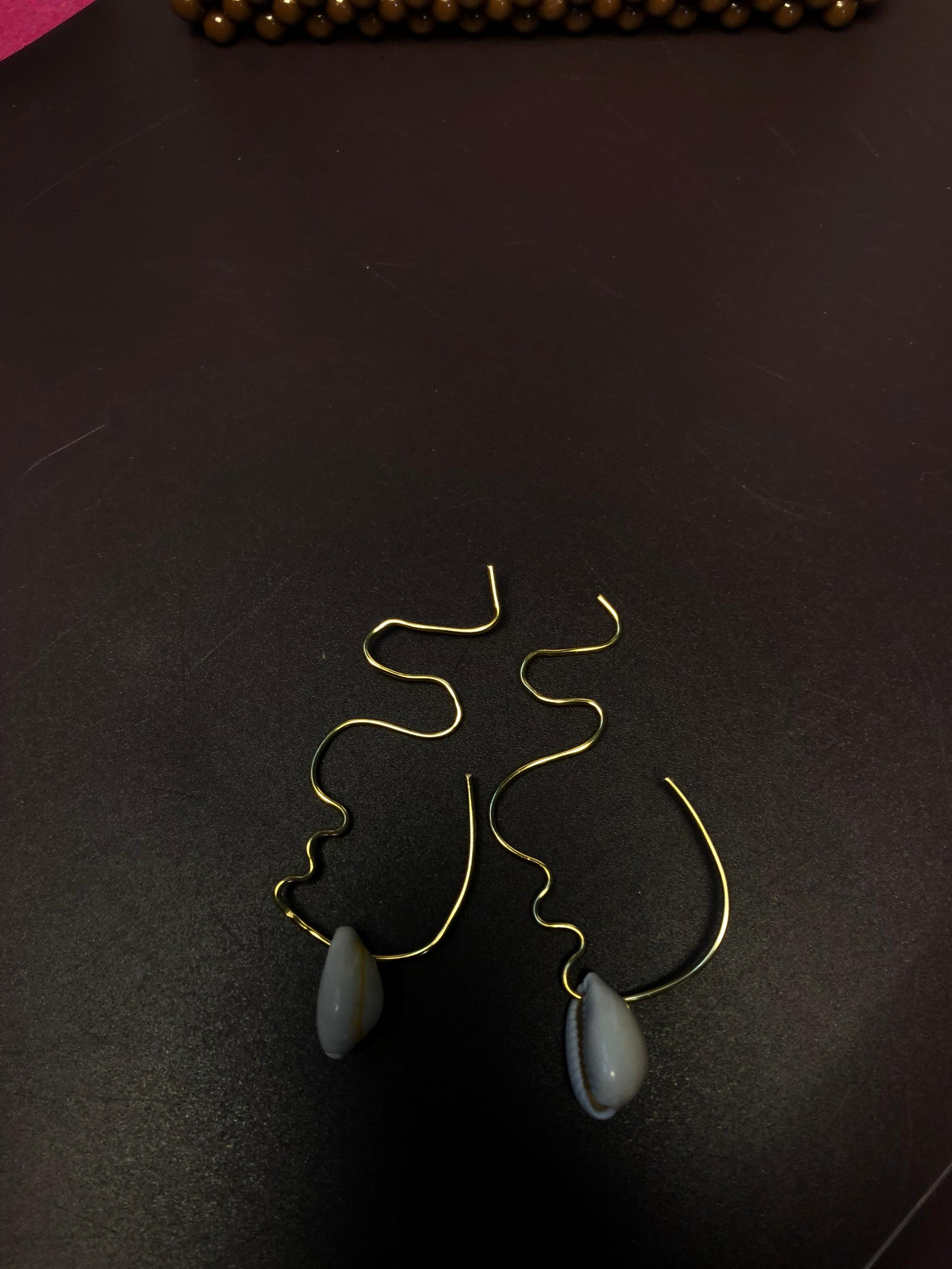 Paire de boucles d'oreilles originales en fil d'or façonnées à la main dessinant le profil abstrait d'un visage avec un cauris naturel suspendu à la base, posées sur un fond sombre.