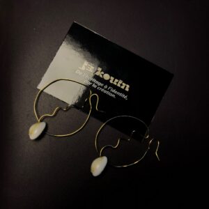 Une paire de boucles d'oreilles créoles originales en fil doré torsadé, chacune ornée d'un coquillage cauri blanc naturel à sa base, posées sur un fond sombre à côté d'une carte jekouin.