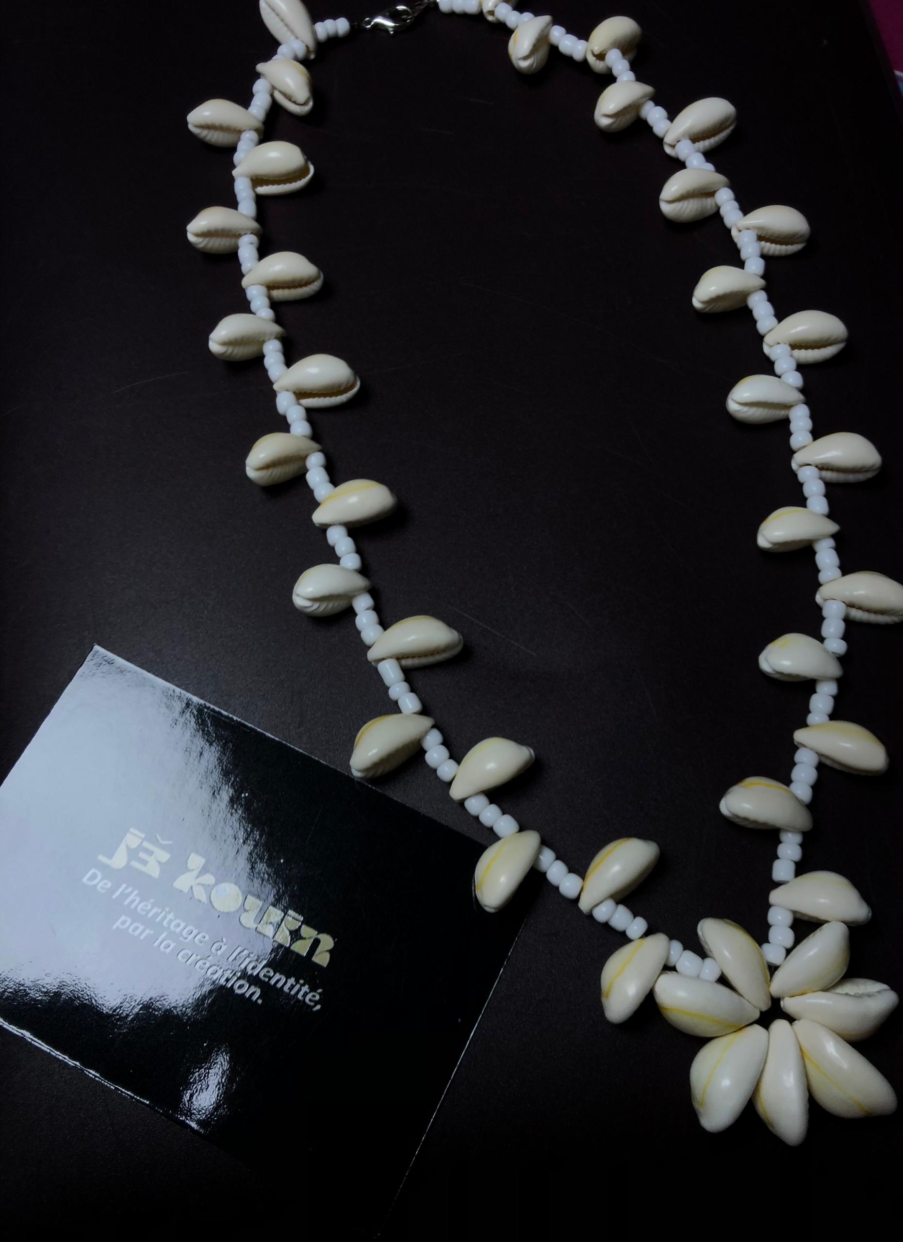 Collier plastron composé d'un rang de cauris et de perles blanches se terminant par un pendentif en forme de fleur de cauris, posé à côté d'une carte jekouin.