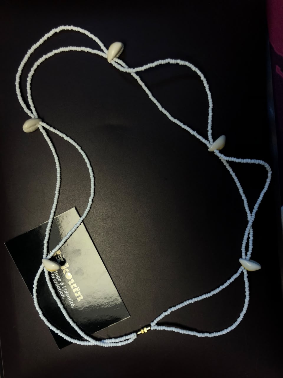 Une parure de tête ou diadème composé de rangs de micro-perles blanches entrelacées et orné de coquillages cauris, disposé sur fond noir avec la carte jekouin.