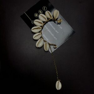 Bijou d'oreille de style ear cuff en forme de croissant de lune orné de cauris blancs naturels avec une longue chaîne dorée suspendue, posé sur fond noir avec la carte jekouin.