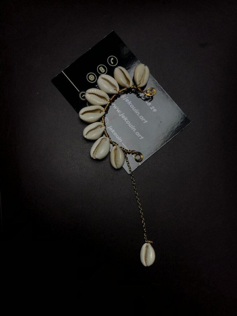 Bijou d'oreille de style ear cuff en forme de croissant de lune orné de cauris blancs naturels avec une longue chaîne dorée suspendue, posé sur fond noir avec la carte jekouin.