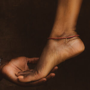 Pied féminin paré d'un bracelet de cheville fin en perles rouges et d'une petite chaînette dorée, délicatement soutenu par une main d'homme sur fond terricole.