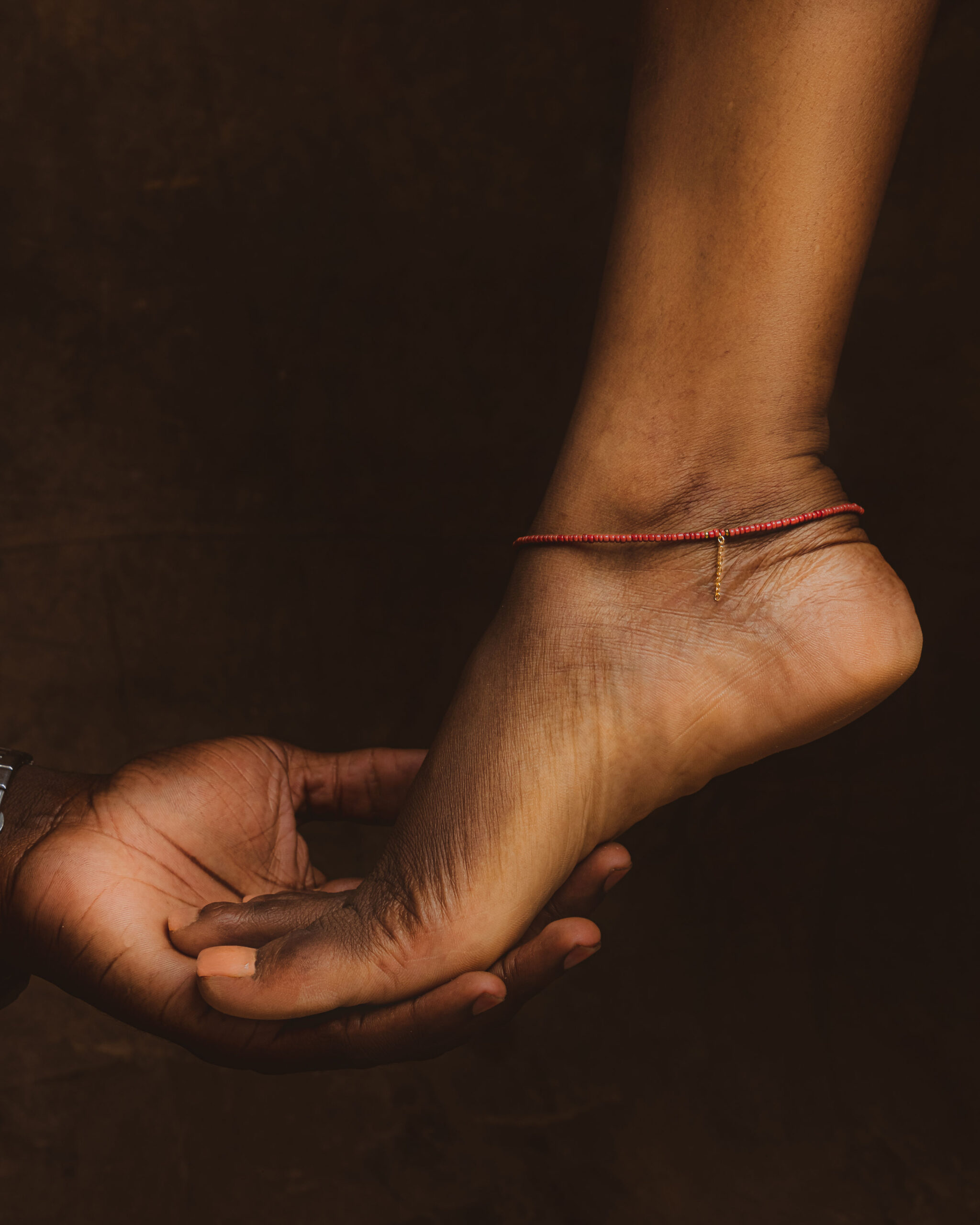 Pied féminin paré d'un bracelet de cheville fin en perles rouges et d'une petite chaînette dorée, délicatement soutenu par une main d'homme sur fond terricole.