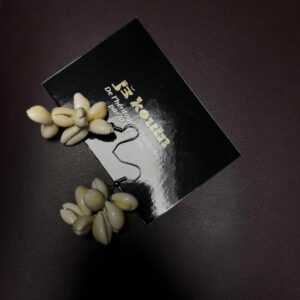 Gros plan sur une paire de boucles d'oreilles pendantes formées de grappes volumineuses de cauris naturels, posées sur une carte de visite jekouin sur fond noir.