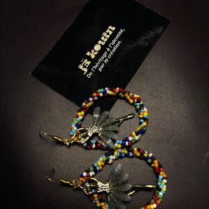 Gros plan vertical sur une paire de boucles d'oreilles créoles ornées de perles multicolores et abritant une fée dorée en leur centre, sous la carte jǯ kouin.