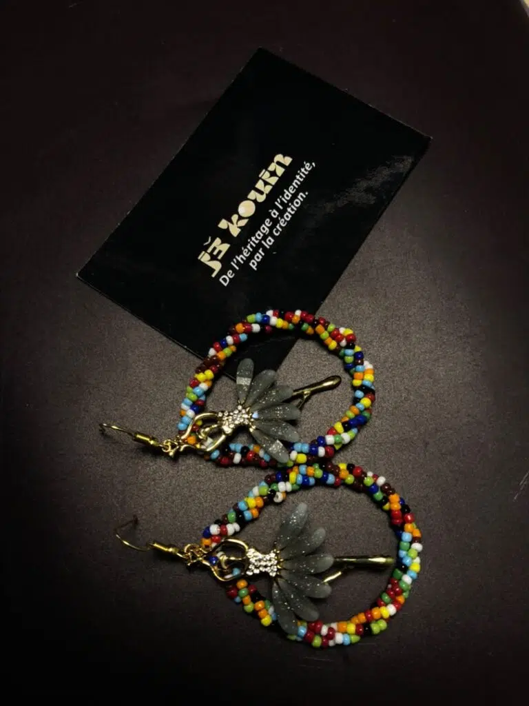 Gros plan vertical sur une paire de boucles d'oreilles créoles ornées de perles multicolores et abritant une fée dorée en leur centre, sous la carte jǯ kouin.